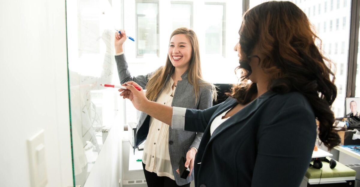 Two professional women discussing ideas on a whiteboard in a modern office setting