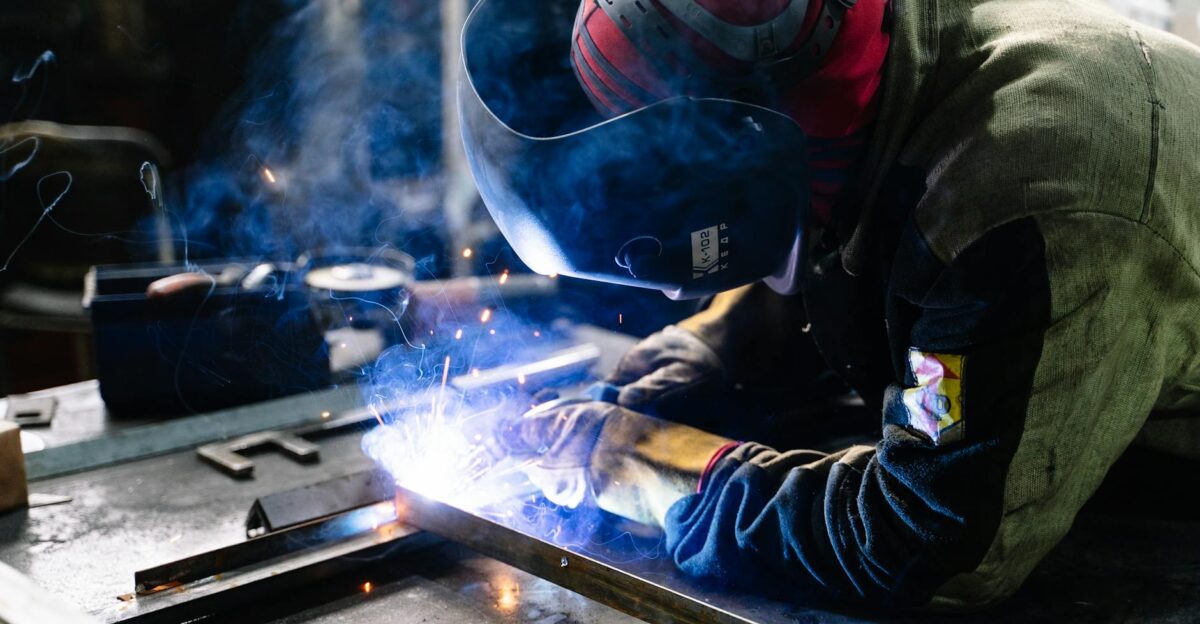 A welder works on metal fabrication indoors creating bright sparks and smoke
