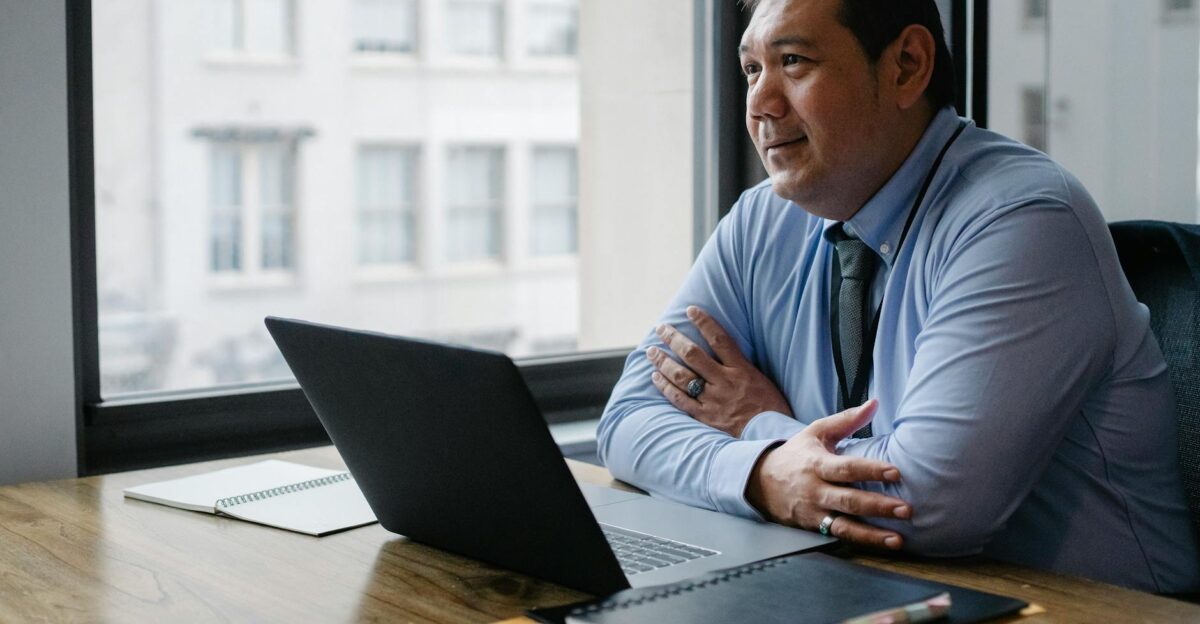 Adult successful ethnic male boss wearing shirt and tie sitting with hands crossed at workplace with documents and netbook