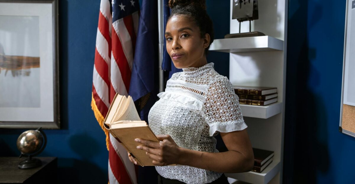 Professional woman holding a book poses confidently in an office setting with flags in the background