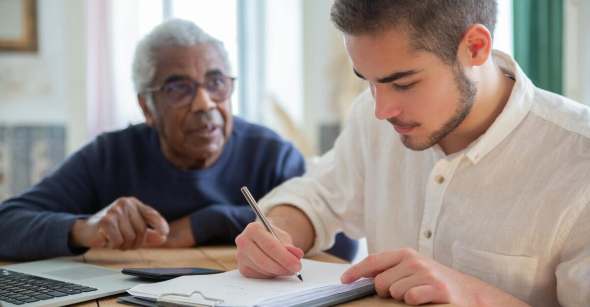 A young man aids a senior citizen with writing at a wooden table indoors fostering social assistance and connection