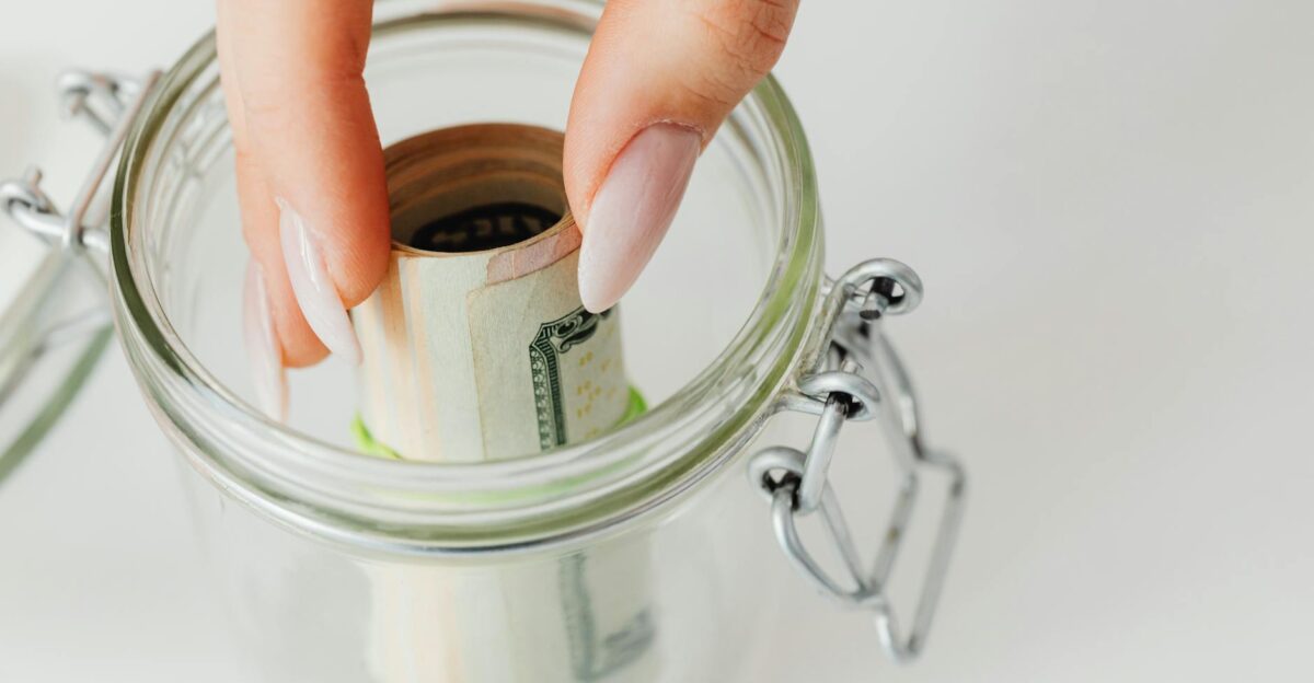 Close-up of hand placing rolled currency into a clear glass jar symbolizing savings