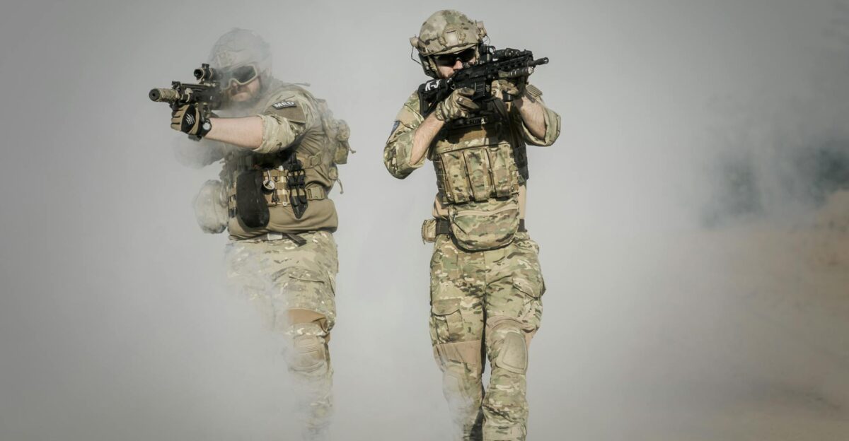 Two soldiers in camo uniforms with weapons advancing through a smoke-covered field showcasing military action