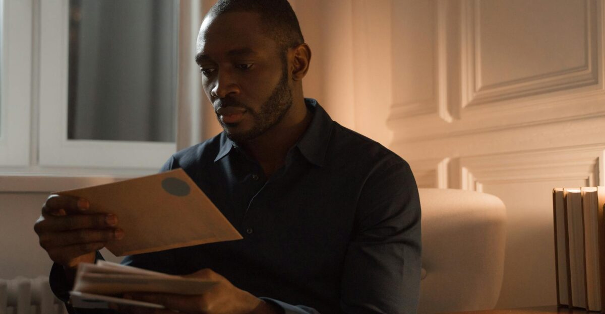 A man with a mustache reads letters while sitting in a warmly lit home interior