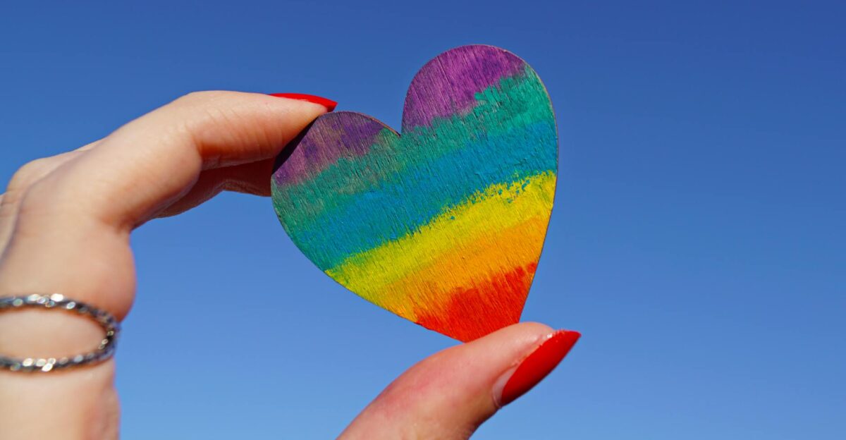A hand holding a rainbow-colored heart against a clear blue sky symbolizing love and pride