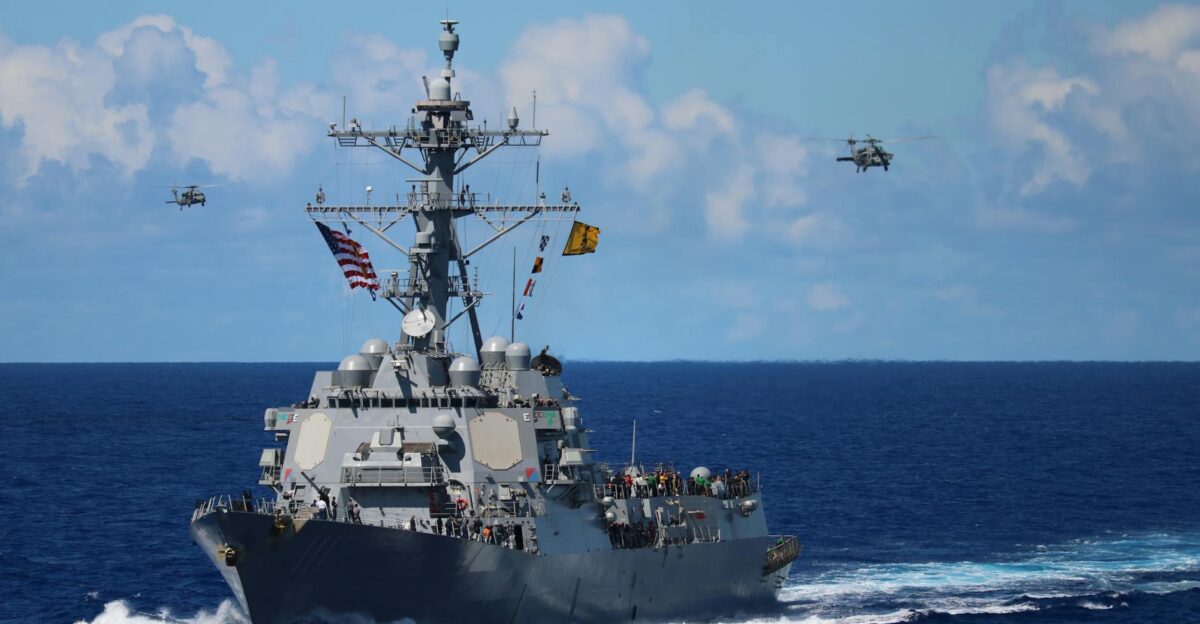 A US Navy destroyer navigates the ocean with helicopters in formation above