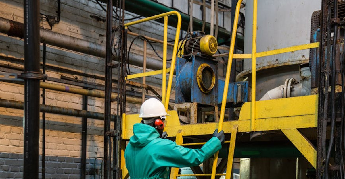 A worker in safety gear climbs a yellow ladder in an industrial factory setting surrounded by machinery