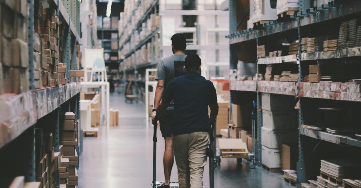 Two men maneuver a trolley in a large warehouse filled with boxes and shelves