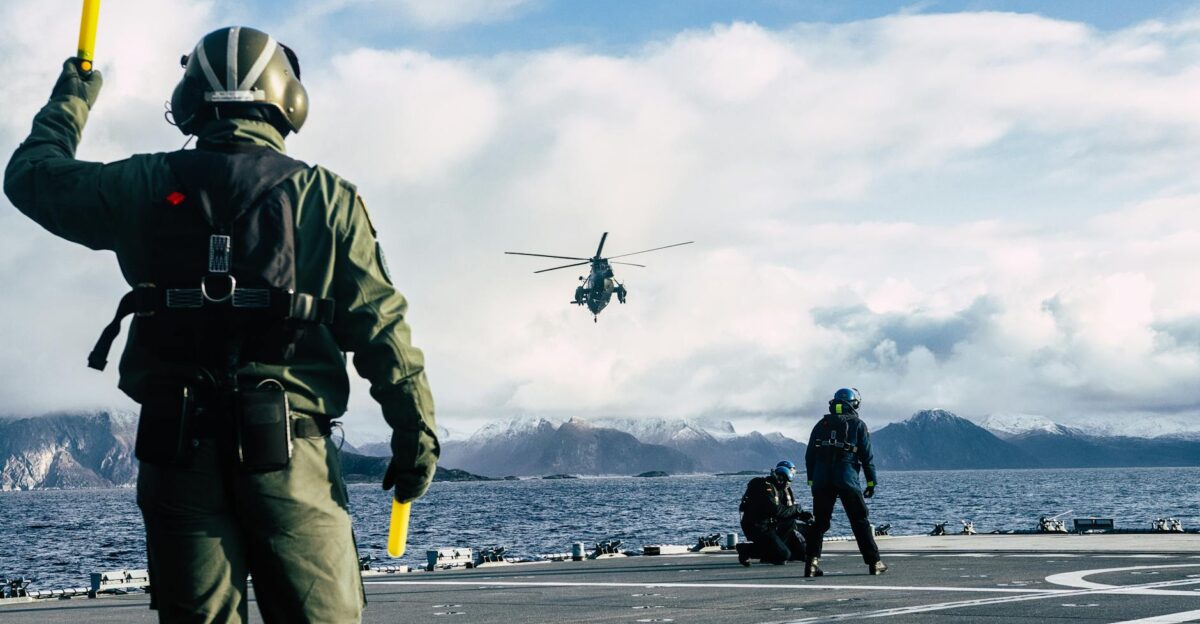 Military personnel directing helicopter landing on a ship s deck with scenic ocean and mountain backdrop