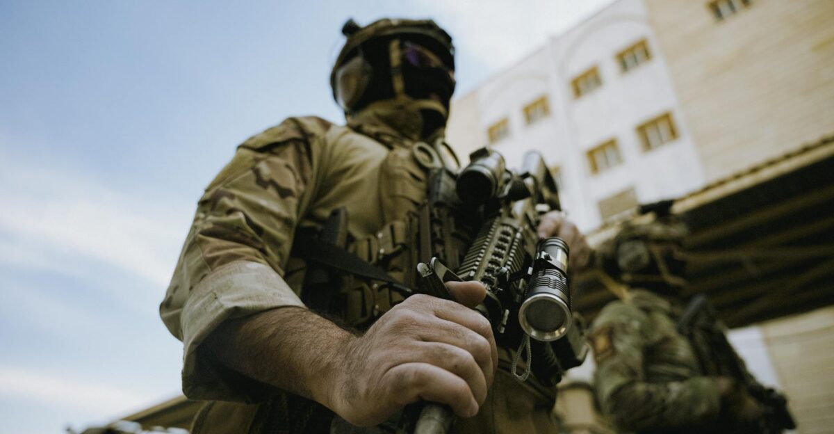 A soldier in tactical gear holding a rifle outdoors by buildings