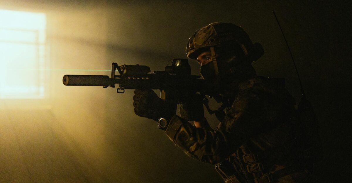 A soldier in tactical gear aims a weapon with a laser sight in low light conditions