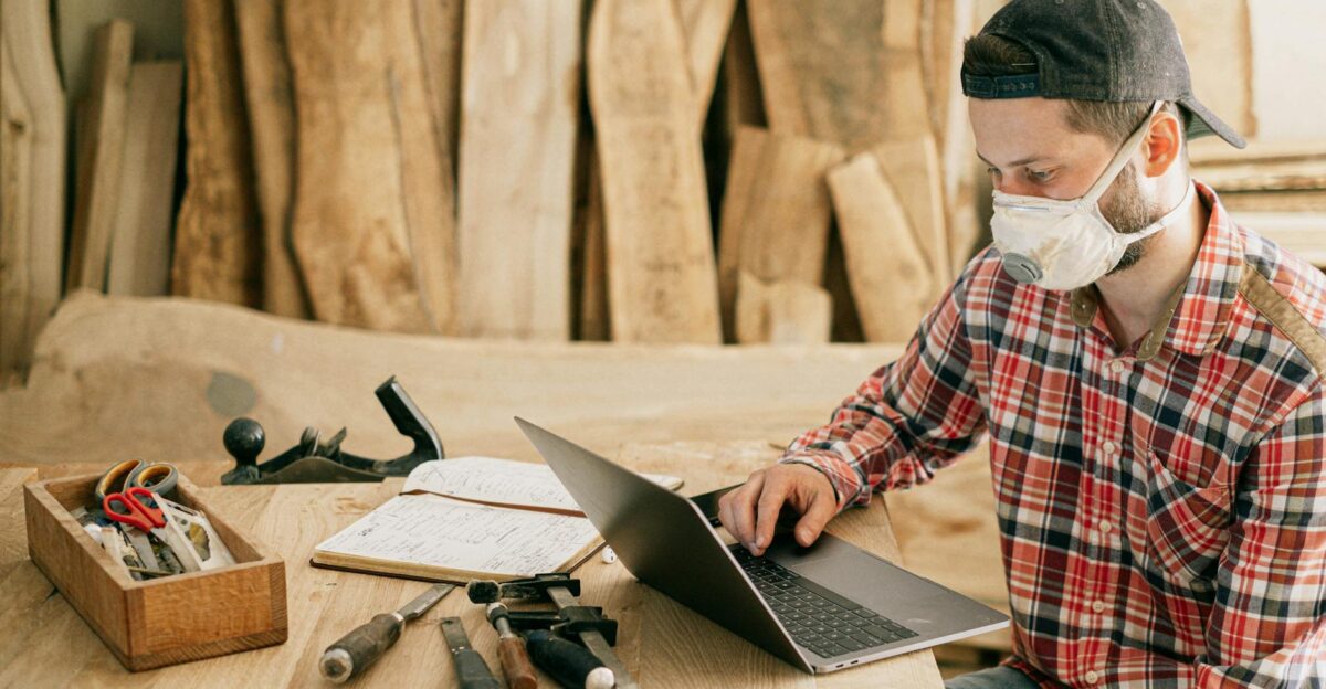 A craftsman wearing a mask working on a laptop in a woodworking shop