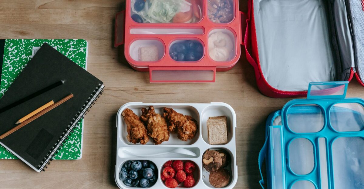 Overhead view of healthy lunchboxes with fruits chicken and notebooks on a wooden desk