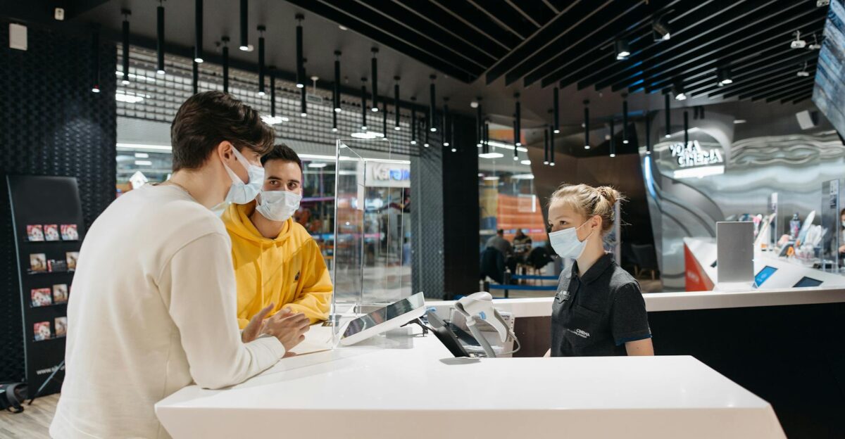 Two masked customers at a modern store counter interacting with a masked female staff member