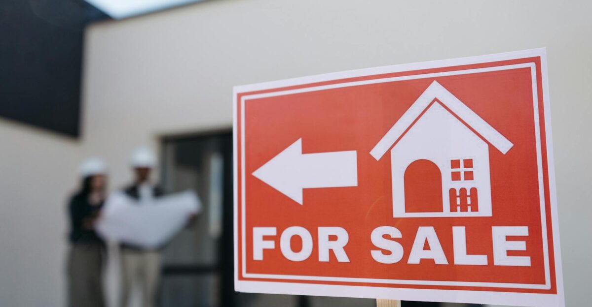 A real estate sign indicates a property for sale as two agents in hard hats discuss building plans outdoors