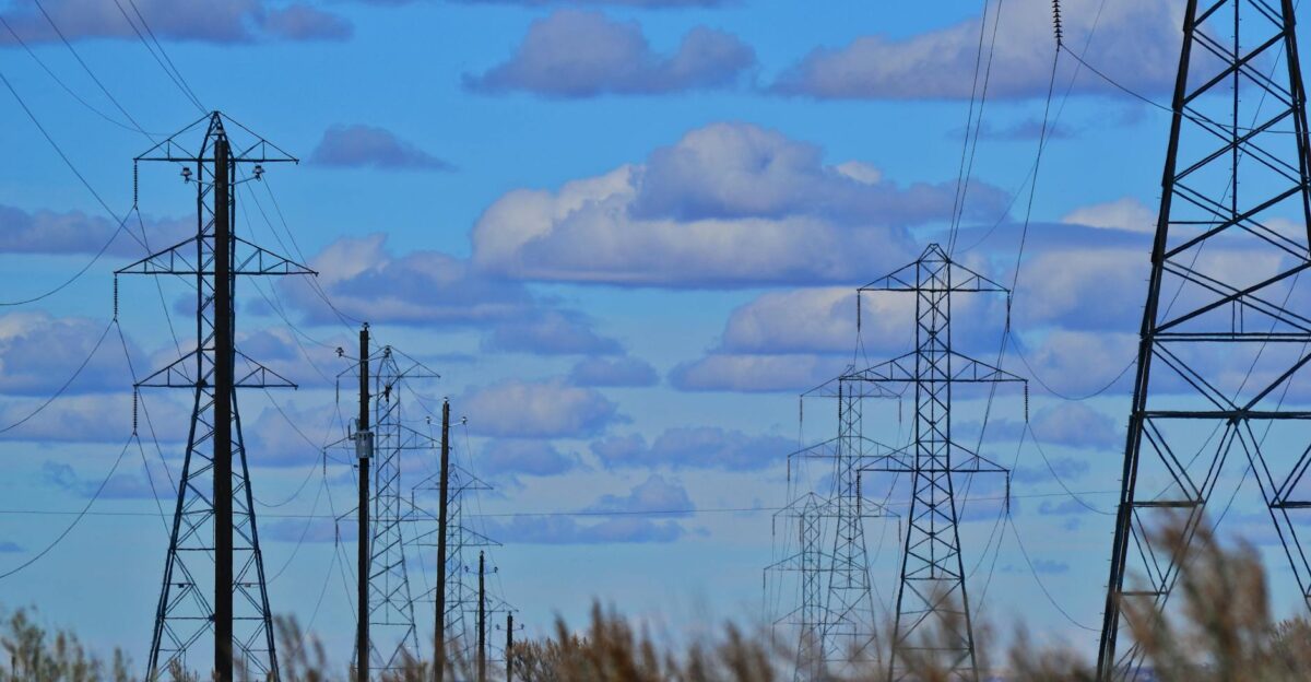 Transmission towers and power lines silhouetted against a bright blue sky with clouds