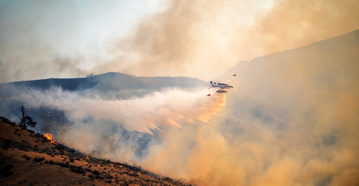 A firefighting plane combats a wildfire spreading water over rugged terrain at sunset