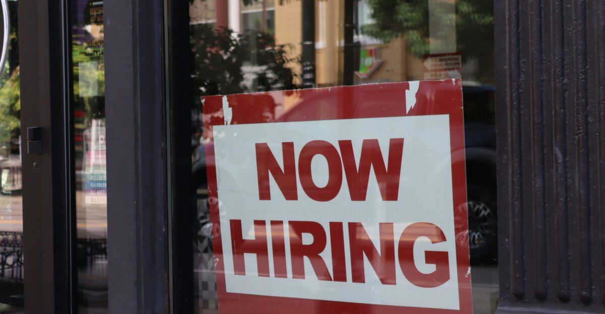 A now hiring sign hangs in a store window