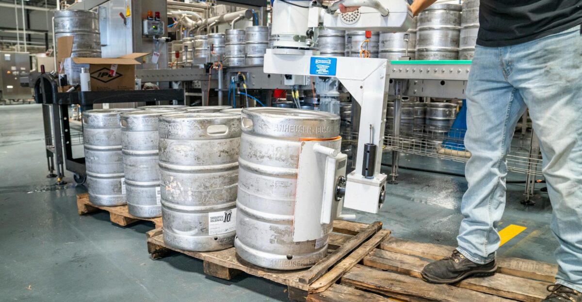A worker operates machinery to stack steel kegs in a brewery warehouse