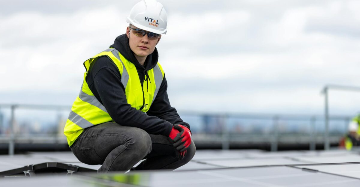 A construction worker inspects rooftop solar panels wearing safety gear and high visibility vest