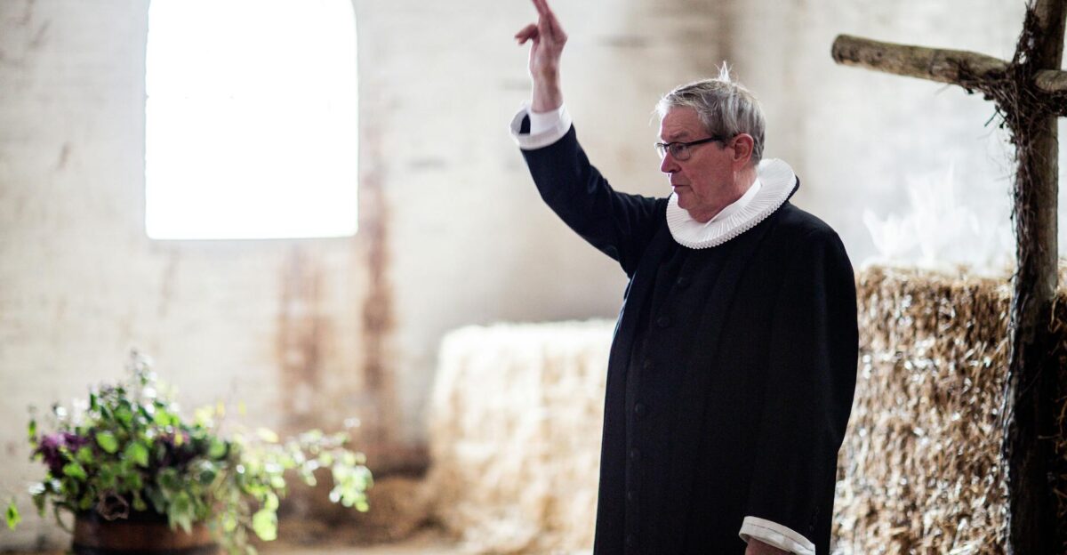 Portrait of an elderly priest conducting a ceremony in a rustic barn setting with hay bales and natural light