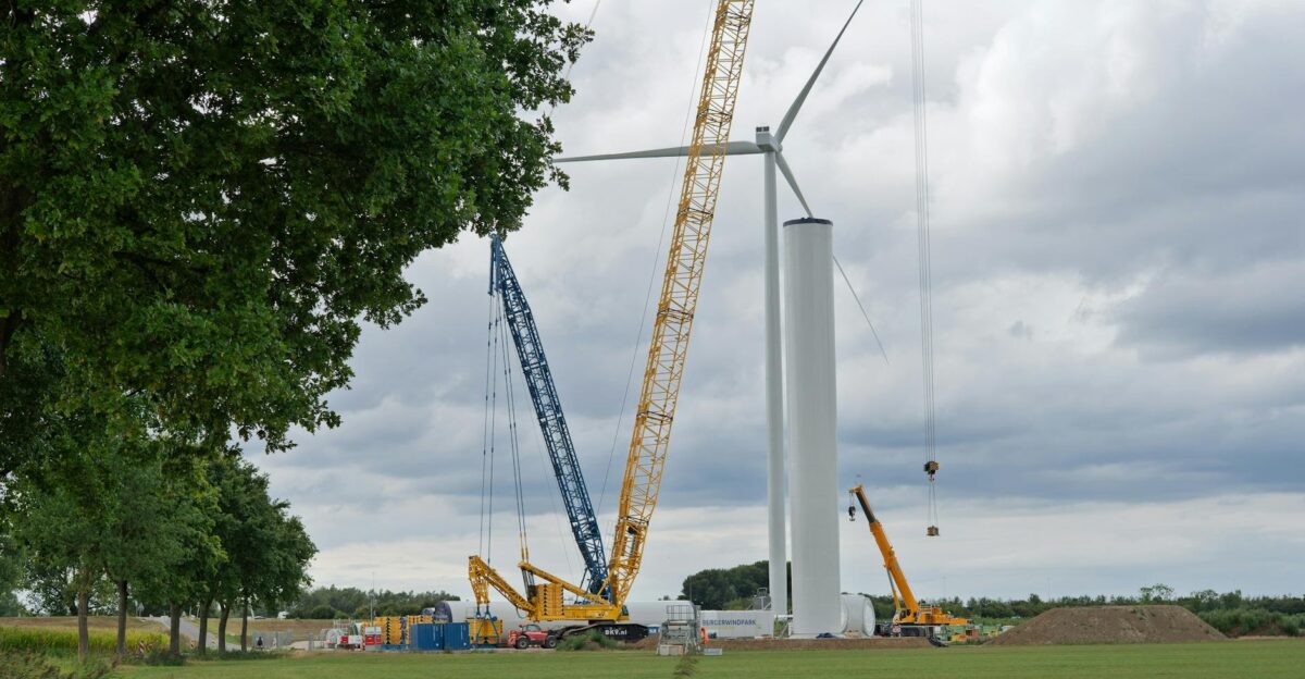 Cranes assembling a wind turbine in a grassy field under a cloudy sky