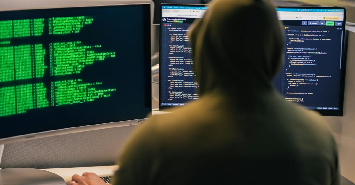 A person in a hoodie coding on dual monitors depicting cybersecurity and hacking themes