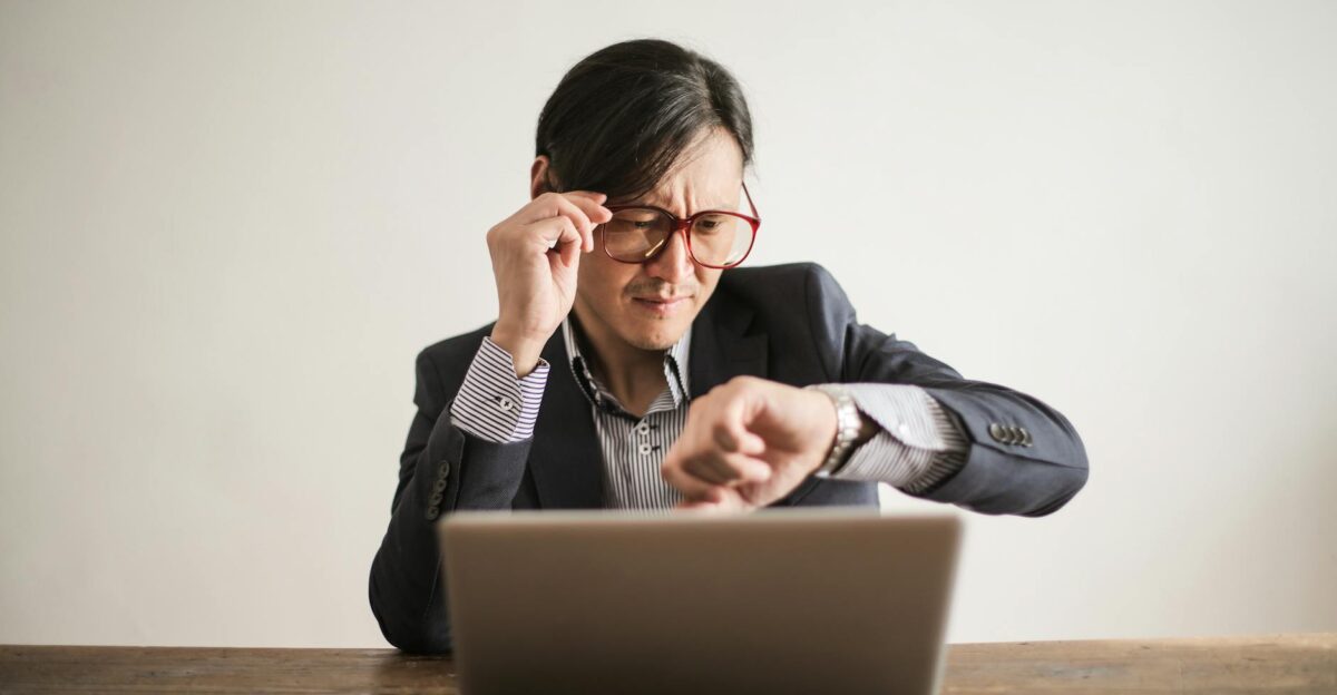 Young frowning man in suit and glasses looking at wristwatch while waiting for appointment sitting at desk with laptop