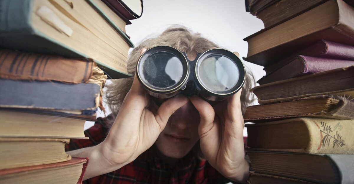 A man observing through binoculars surrounded by stacks of vintage books indoors