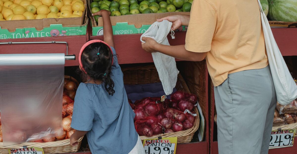 Back view of crop anonymous mother with reusable bag and girl choosing limes from box