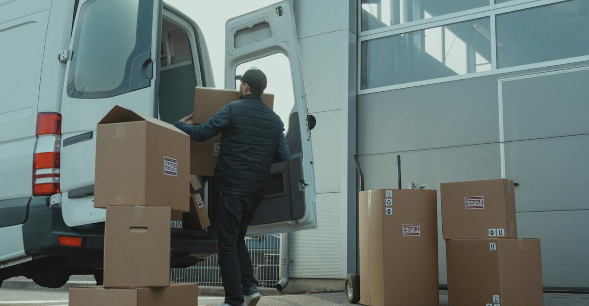 A delivery man unloading cardboard boxes from a van at a warehouse during the day