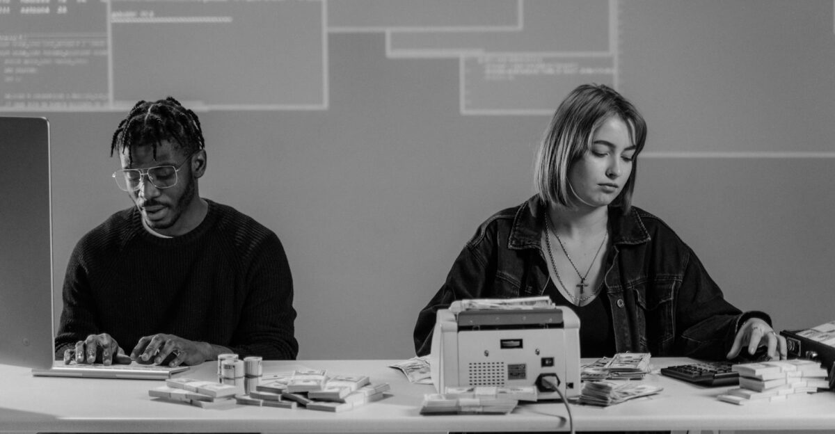 Two professionals working with computers and currency at a desk in a modern office setting