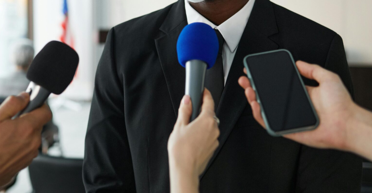 African American businessman in formal attire speaks to media while wearing a face mask in a conference room setting.