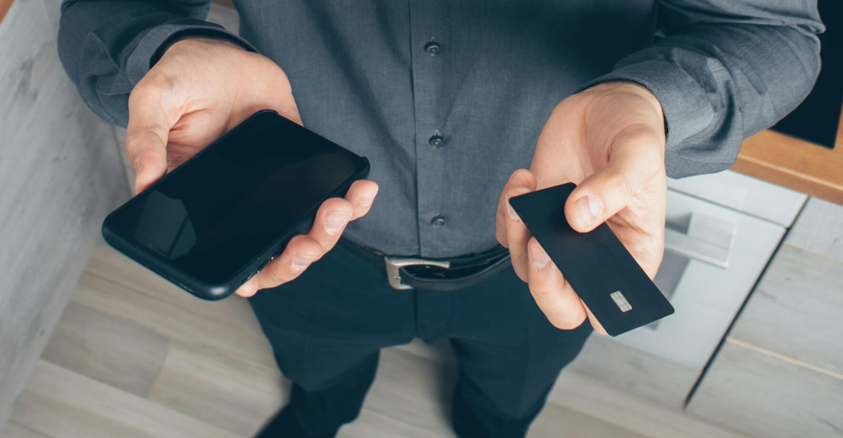 A confused man indoors looks up while holding a smartphone and credit card, concerned about a financial issue.