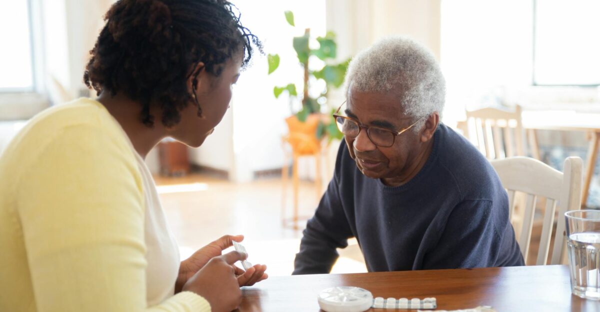 A woman and an elderly man discuss medication at a wooden table in a bright room highlighting care