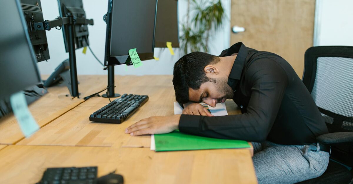 An exhausted office worker resting on a desk surrounded by computer monitors in a modern workspace