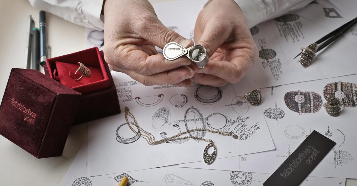 Close-up of hands inspecting a diamond ring with a loupe on a workbench filled with jewelry sketches