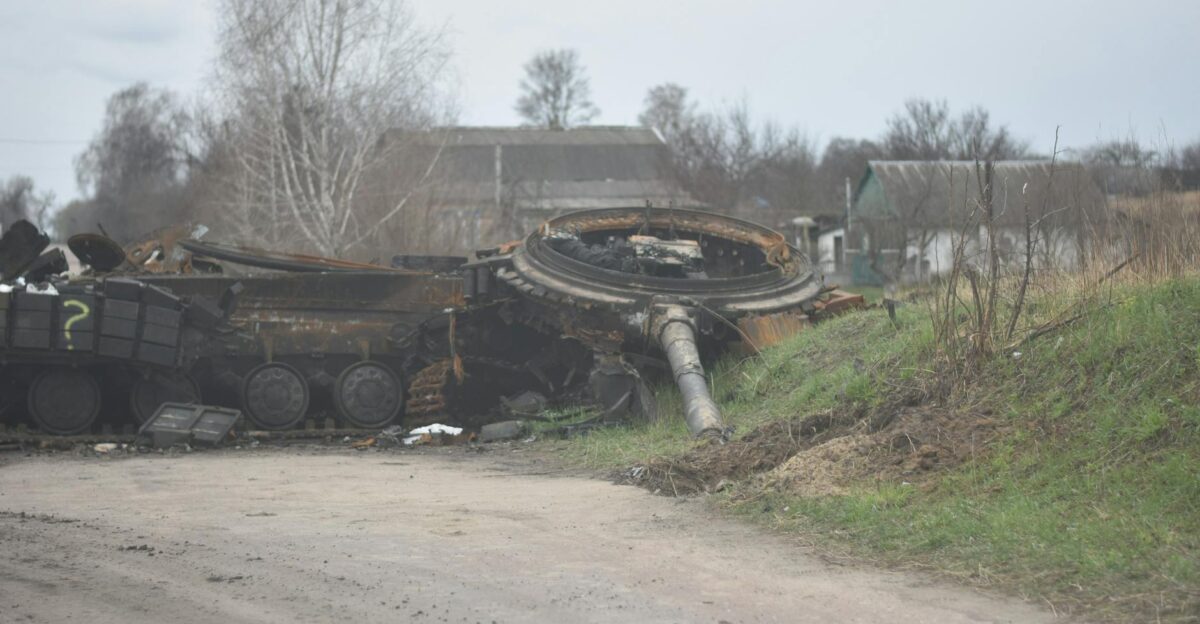 A destroyed military tank wreck on a dirt road near rural houses