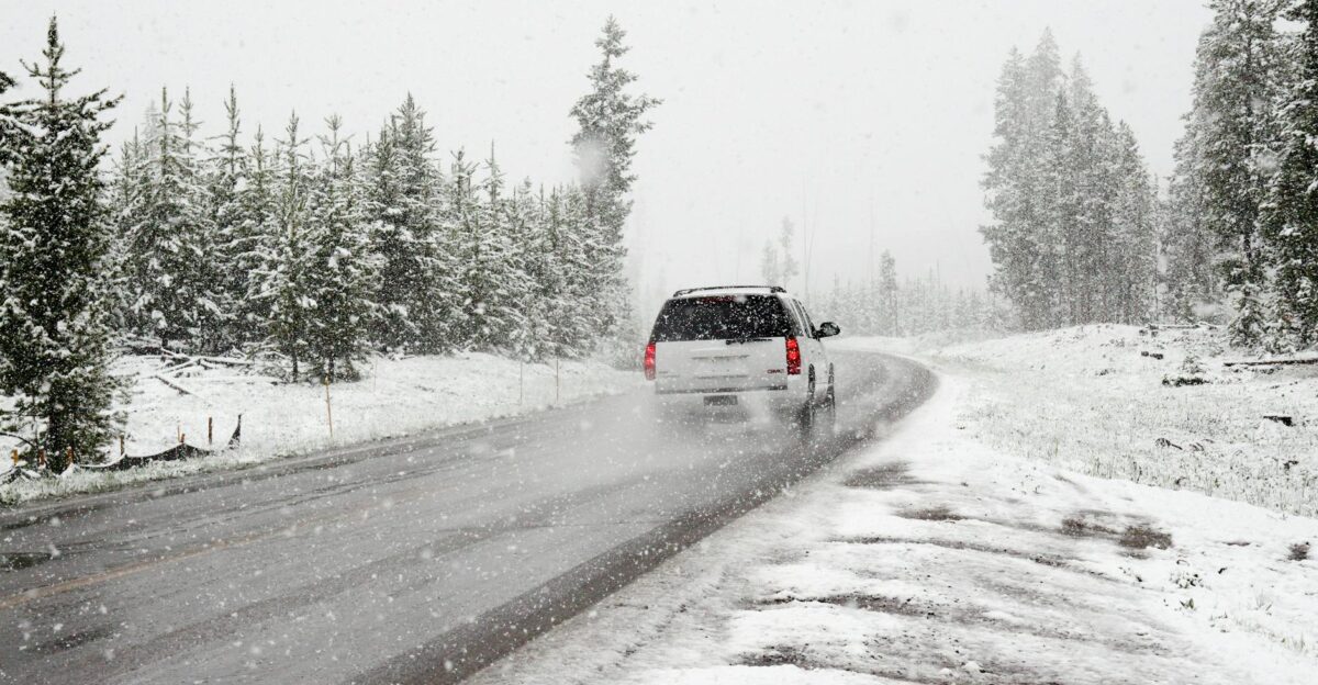A car drives on a snowy road surrounded by trees during a snowstorm creating a wintry scene