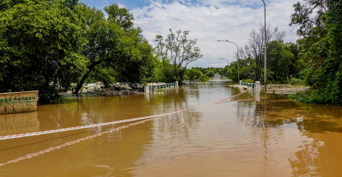 A flooded road with a submerged bridge surrounded by trees under a cloudy sky
