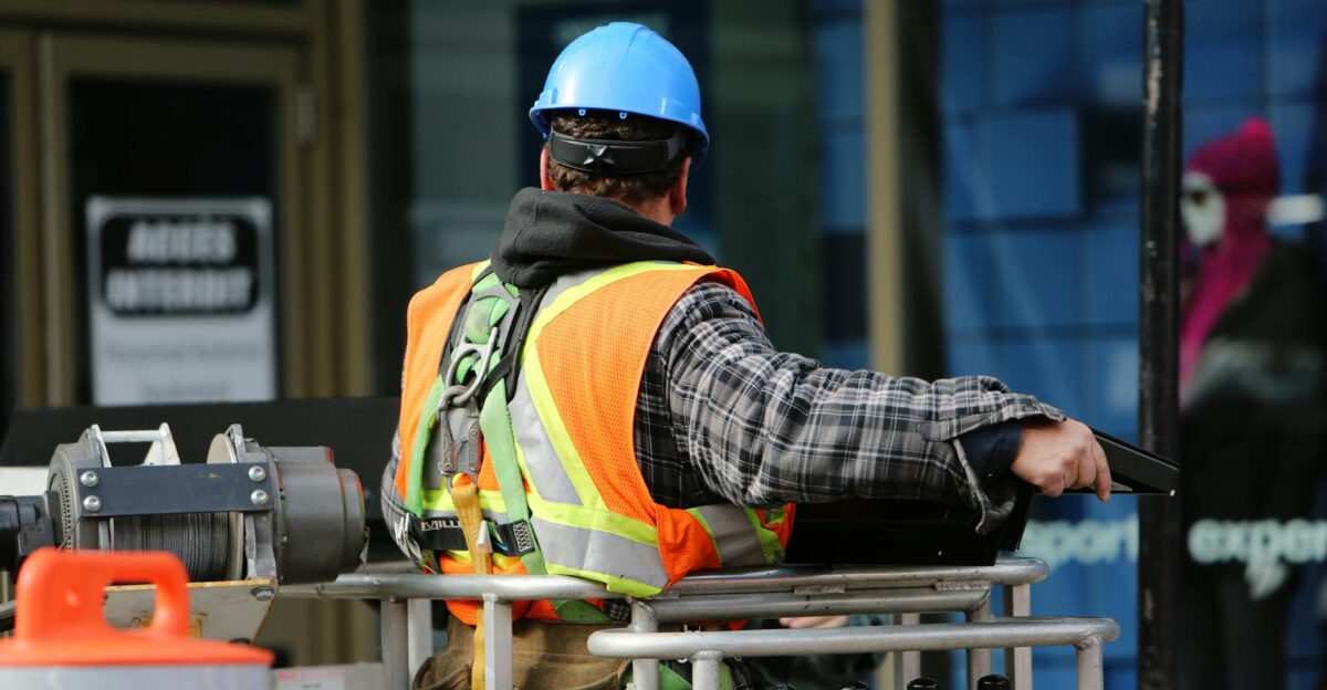 A construction worker wearing a blue helmet and safety vest operates equipment at an urban construction site