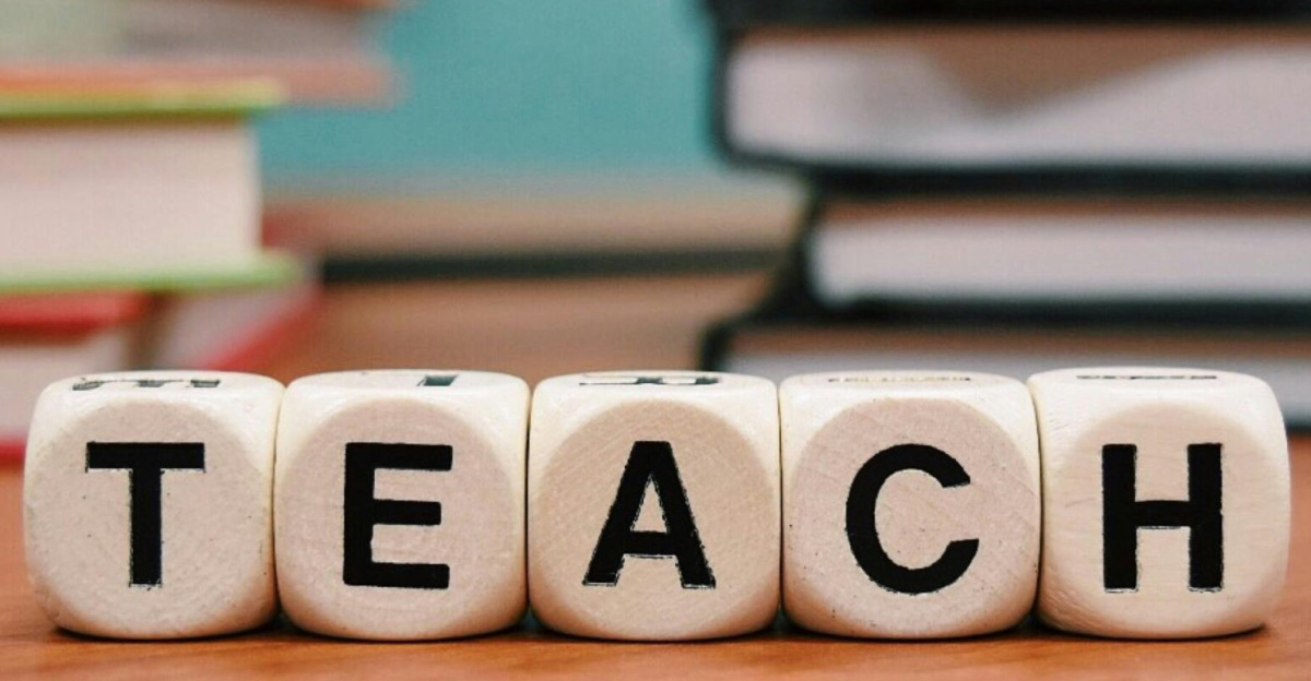 Close-up of dice spelling 'TEACH' amidst stacked books on a wooden table.