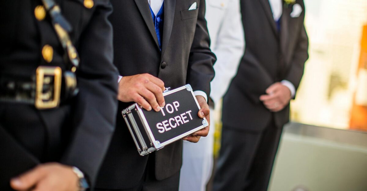 A group of men in formal military and business attire holding a Top Secret briefcase indoors