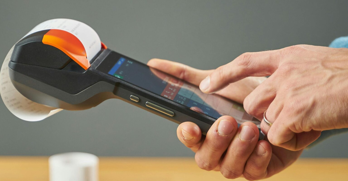 Hands operating a portable receipt printer with printed receipts on a wooden table.