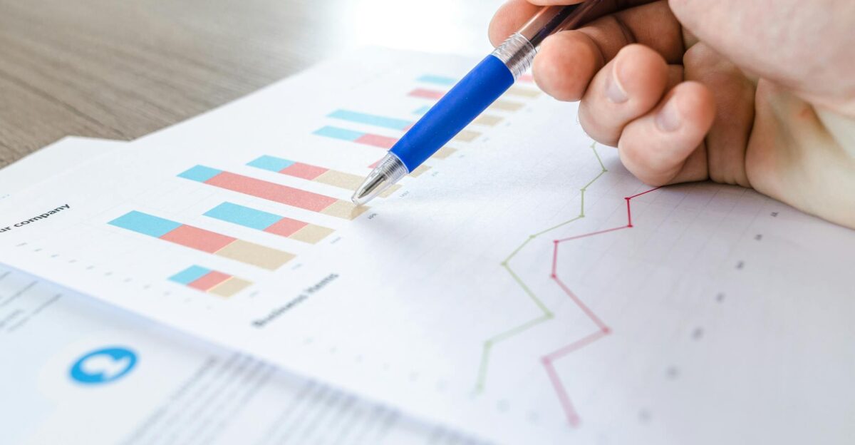 A close-up of a hand with a pen analyzing data on colorful bar and line charts on paper