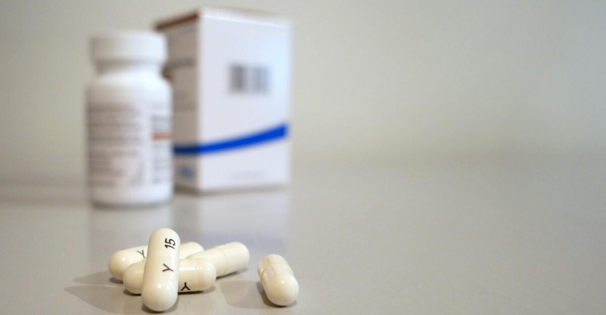 Close-up image of white capsules with medicine bottle and box in the background