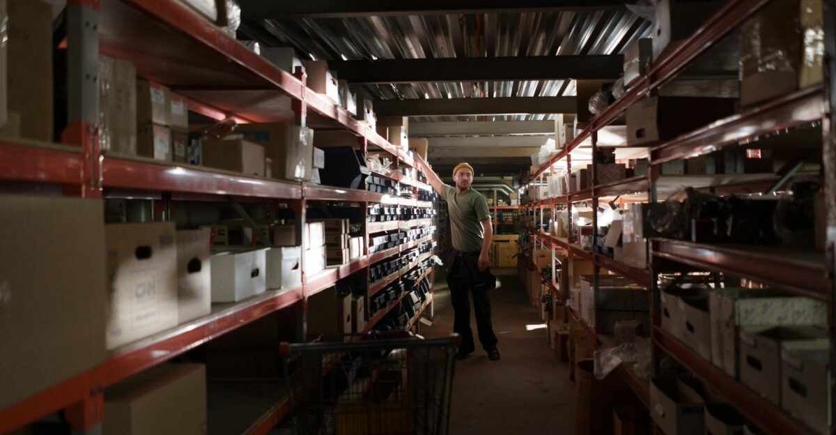 A worker organizing inventory in a dimly lit warehouse aisle full of shelves and boxes
