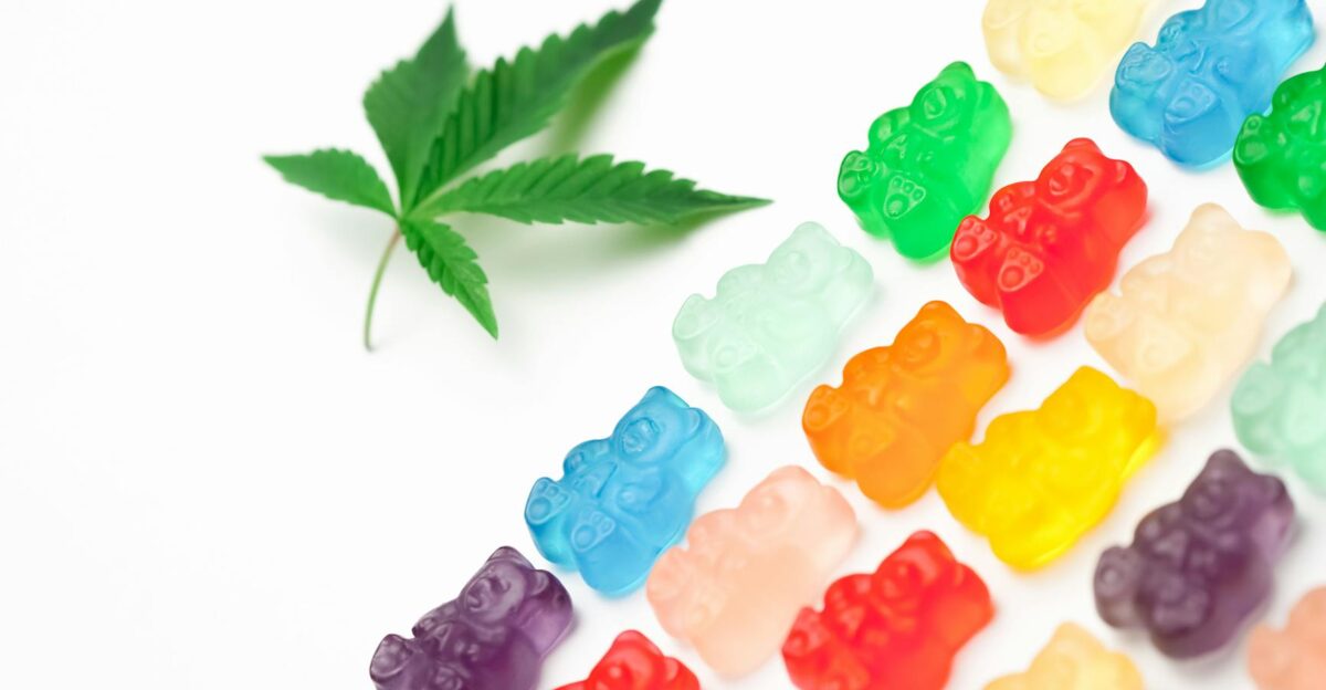 Vibrant gummy bears next to a cannabis leaf on a white background symbolizing edible cannabis sweets
