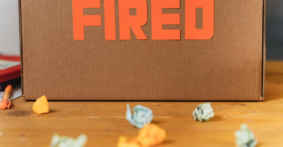 Close-up of a cardboard box labeled FIRED on a wooden table with crumpled papers