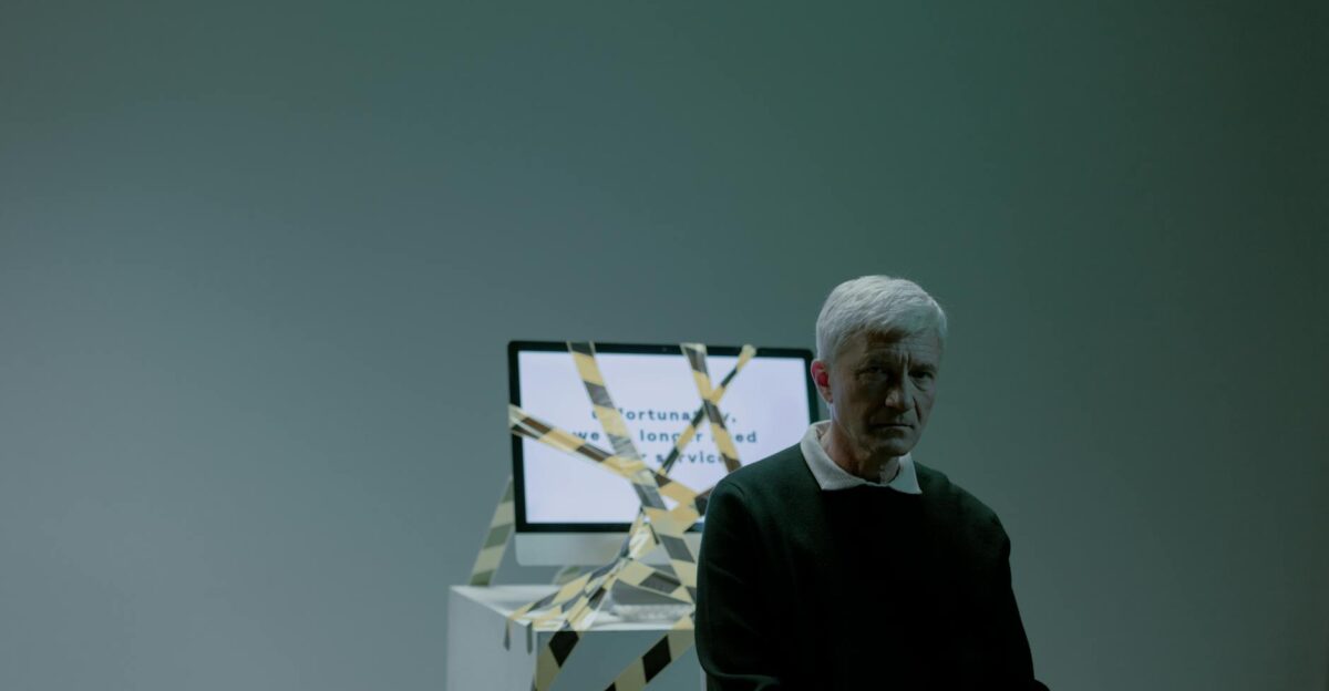 A senior adult sits indoors next to a computer wrapped in caution tape symbolizing job loss and unemployment